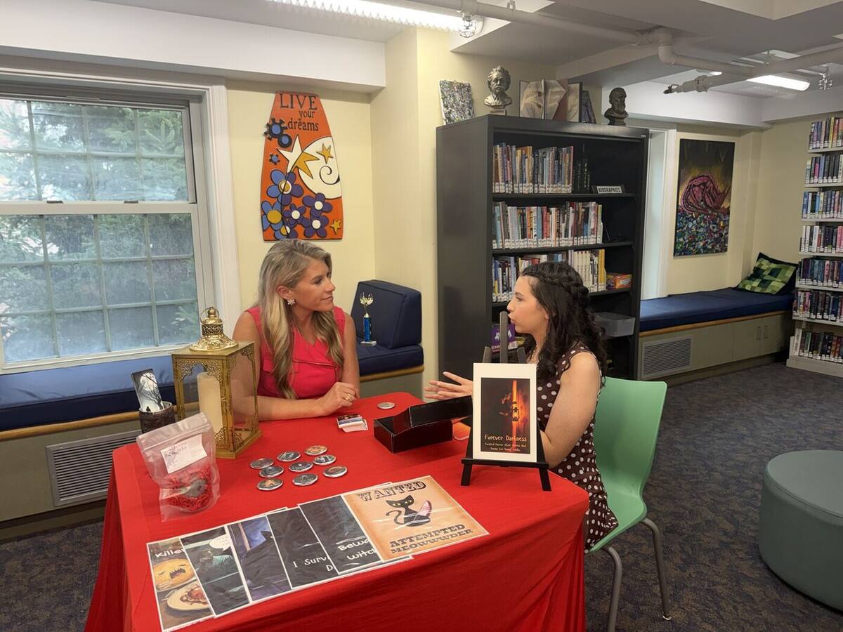 Ma'ayan being interviewed at the Forever Darkness display table, with pins, cards, and the book on show